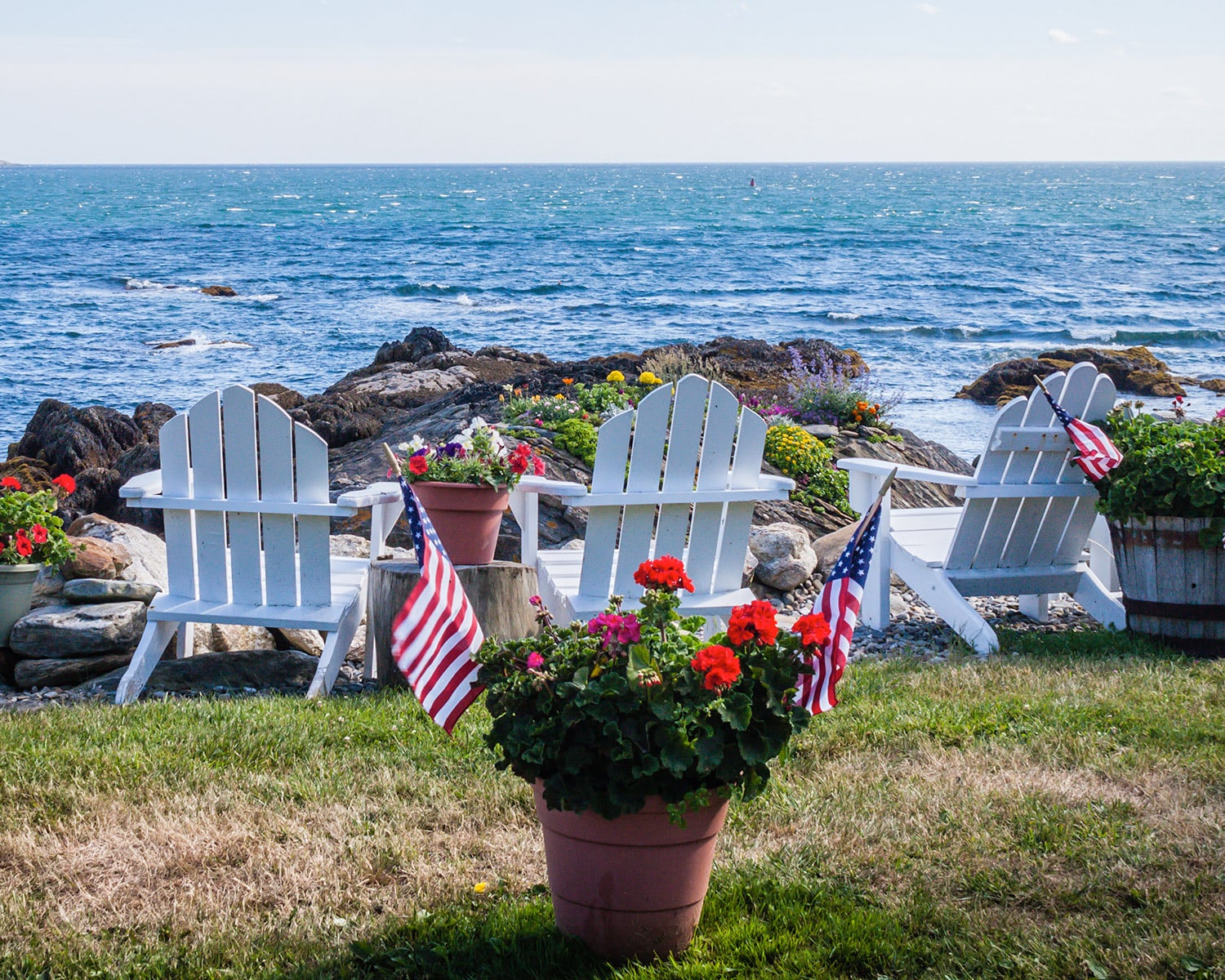 Coastal patio with American flags, red geranium plants, and white wooden chairs overlooking the ocean